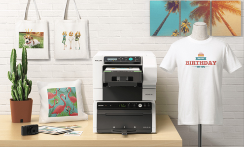 RICOH Ri 100 mit Textil-Finisher auf einem Holztisch, umgeben von bedruckten Textilien wie T-Shirts, Kissen und Stofftaschen – kompakter DTG-Drucker für kreative On-Demand-Produktion