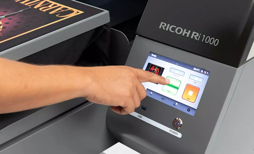 Finger tapping the touchscreen of the RICOH Ri 1000X while a black T-shirt with colorful print lies on the platen – intuitive DTG printer for individual textile designs