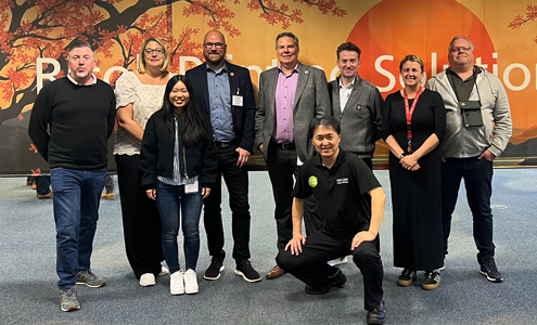 Group photo of nine people in front of a RICOH Textile Solutions wall graphic with red sun and cherry blossom design – international team at a textile printing solutions event