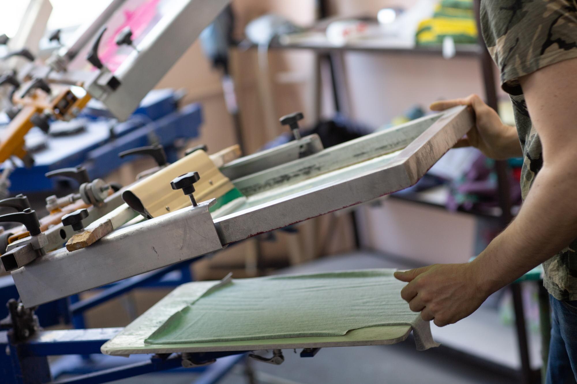 A person lifts a screen printing frame over a green garment placed on a printing pallet in a workshop.